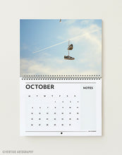 Load image into Gallery viewer, October page of the 2027 A3 art wall calendar, pair of trainers hanging from a wire in a blue sky with a plane trail, minimalist surreal photography
